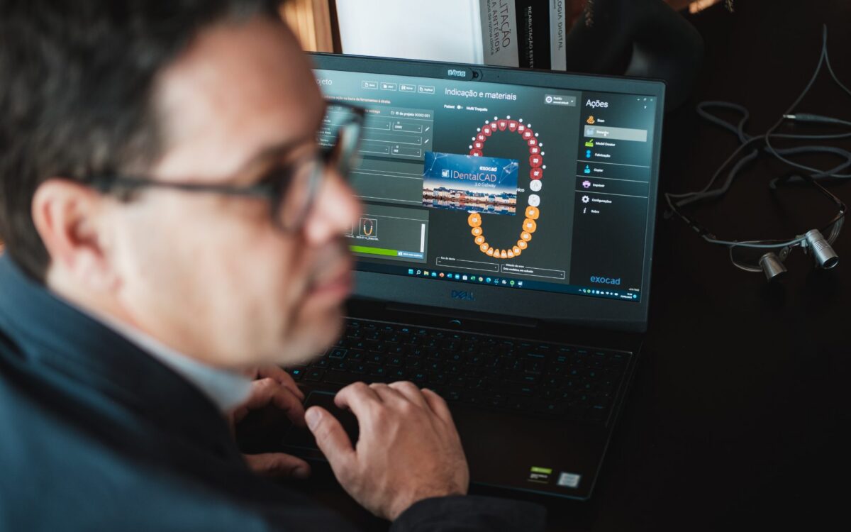 Homem de óculos e camisa azul usando laptop Dell com software de design odontológico mostrando modelo de dentes em 3D em tela de trabalho