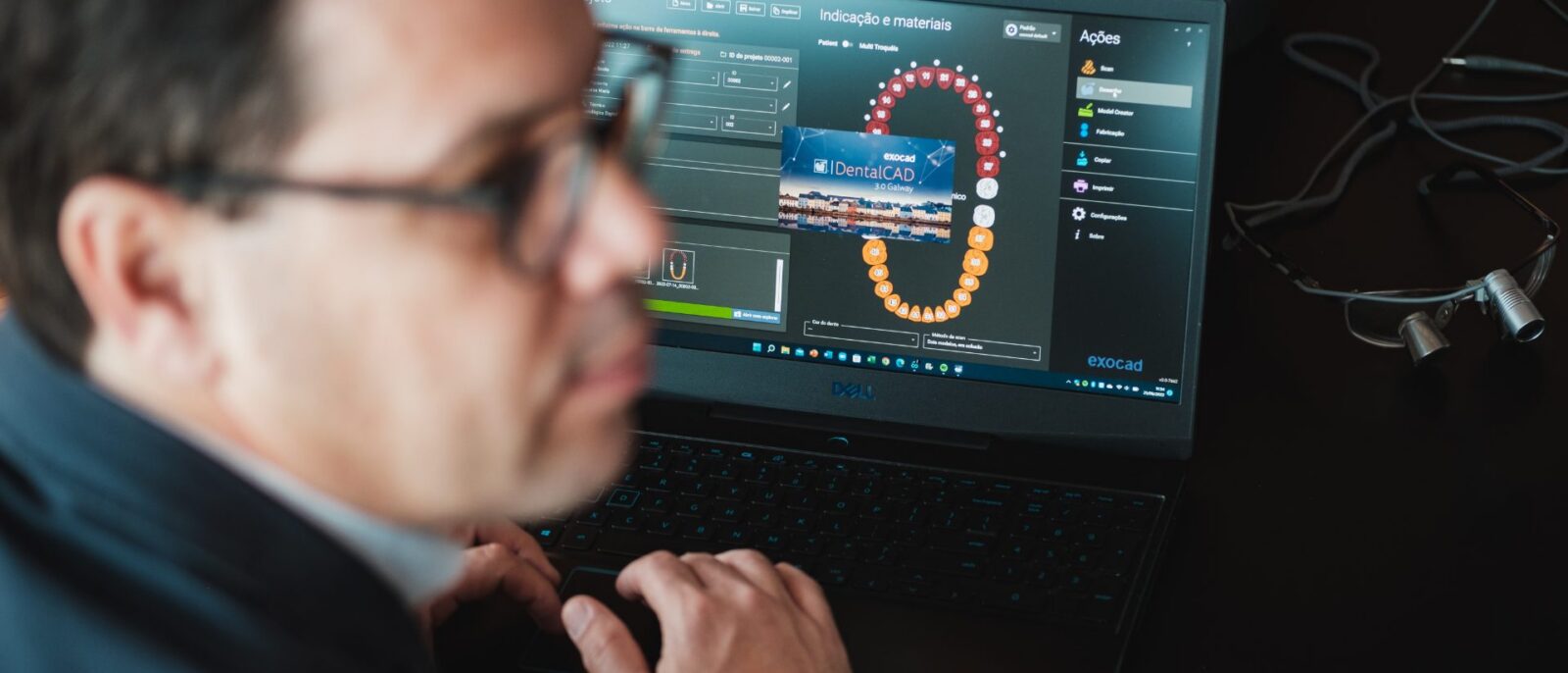 Homem de óculos e camisa azul usando laptop Dell com software de design odontológico mostrando modelo de dentes em 3D em tela de trabalho